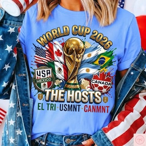 Pode incluir: Uma t-shirt azul-clara com um gráfico da Copa do Mundo de 2026. O design inclui o troféu da FIFA, as bandeiras dos EUA, Canadá e México, e o texto "WORLD CUP 2026", "THE HOSTS", "EL TRI", "USMNT" e "CANMNT" em letras em negrito.