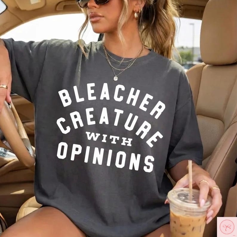 May include: Dark gray t-shirt with the text "BLEACHER CREATURE WITH OPINIONS" in white block letters. The shirt is a casual style, with a relaxed fit. The image shows a person wearing the shirt.