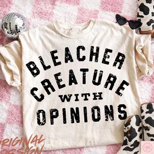 May include: Cream-colored t-shirt with the text "Bleacher Creature With Opinions" in distressed black lettering. The shirt is styled with sunglasses, a disco ball, and cow-print boots.