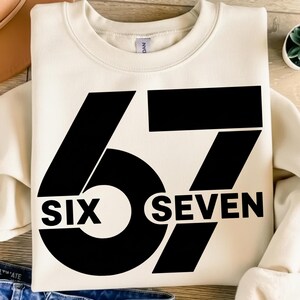 Pode incluir: Camisola creme com o número "67" em letras grandes pretas. A palavra "SIX" está abaixo do "6" e a palavra "SEVEN" abaixo do "7". A camisola é feita de um material macio.