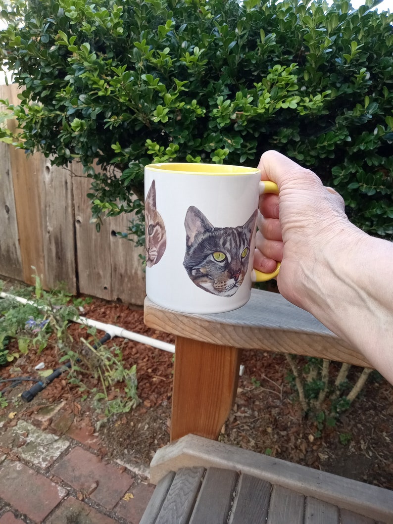 White mug with yellow interior and yellow handle held in right hand above wood arm chair outdoors. Garden plants, red brick path and wood fence in background. View of mug shows partial view of orange cat head and full view of tabby cat head.