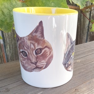 White mug with yellow interior and yellow handle on wood railing outdoors. Leaves in bckground. View of mug shows partial view of tabby cat head and full view of orange cat head.