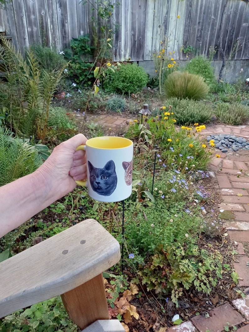 White mug with yellow interior and yellow handle held above wood arm chair in outdoor setting. Garden plants, red brick path and wood fence in background. View of mug shows partial view of orange cat head and full view of russian blue cat head.