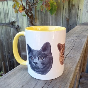 White mug with yellow interior and yellow handle on wood railing in outdoor setting. Leaves and wood fence in background. Photo shows partial view of orange cat head and full view of russian blue cat head.