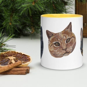 White mug with yellow interior on white tabletop. Greenery in background, Cinnamon and dried orange slices piled next to mug. Photo shows view of orange cat head.
