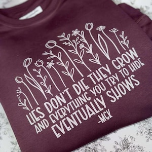 Puede incluir: Sudadera burdeos de cuello redondo con diseño floral blanco y texto: "Lies don't die they grow and everything you try to hide eventually shows -MGK". La sudadera está doblada sobre una superficie con estampado floral.