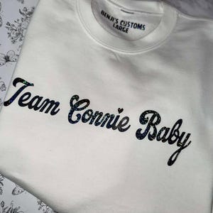 Peut inclure: Un sweat-shirt blanc avec l'inscription "Team Connie Baby" en lettres pailletées bleu foncé. La marque "Benji's Customs" et la taille "Large" sont imprimées sur le col. Le sweat-shirt est en matière douce.