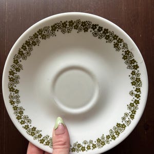 以下が含まれることがあります： 縁に緑色の花柄が施された白い受け皿。受け皿は陶磁器製で、滑らかで光沢のある仕上げです。