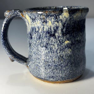 May include: A ceramic mug with a speckled glaze featuring shades of deep navy blue, white, and pale yellow. The mug has a thick, curved handle and a textured, mottled surface finish with a visible brown clay base at the bottom rim.