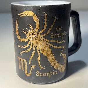 May include: A black ceramic mug featuring a gold-colored illustration of a scorpion. The text "the Scorpion" and "Scorpio" are printed in gold, along with the Scorpio zodiac symbol. The mug has a white interior and a black handle.