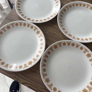 Puede incluir: Cuatro platos de cerámica blanca con un patrón floral dorado alrededor del borde. Los platos están dispuestos sobre una superficie de madera. El diseño floral es un patrón repetitivo de pequeñas flores y hojas.
