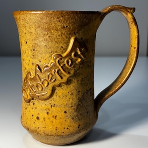 May include: A mustard yellow ceramic mug with a speckled texture and a large handle. The word Oktoberfest is embossed in a raised, stylized script across the front of the mug. The surface has a matte finish with dark brown speckles.
