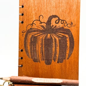 Peut inclure: Carnet en bois avec un motif de citrouille gravé sur la couverture. La citrouille est faite de livres. Un crayon avec le texte "MIDNIGHT ETCH" repose sur le carnet. Le carnet est relié avec du fil de cuivre.