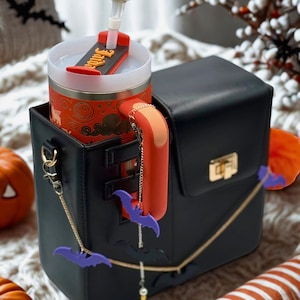 May include: A black handbag with gold detailing, a red handle, and purple bat charms. Inside is a Halloween-themed tumbler with a ghost topper. The tumbler features an orange and black design. Pumpkins and decorations complete the scene.