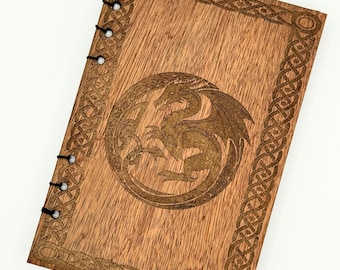 May include: Wooden journal with a dragon design. The cover is made of light brown wood and features a detailed dragon illustration in a circular frame. The border has an intricate Celtic knot design. The journal is bound with black string.