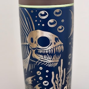 Puede incluir: Una taza de viaje de acero inoxidable azul con un diseño metálico dorado de esqueletos de peces y burbujas. La taza tiene una tapa marrón y la palabra "owala" está impresa en el lateral.