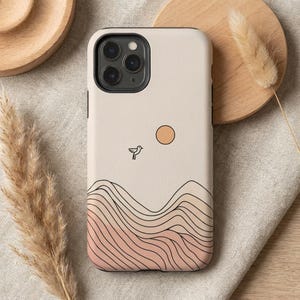 May include: A beige phone case featuring a minimalist landscape design. The case has a black border and a graphic of a bird, sun, and layered mountain range in shades of pink and orange.