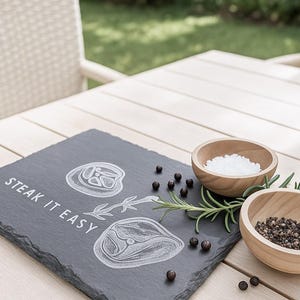 May include: A rectangular slate serving board with the words "STEAK IT EASY" and steak illustrations. Two small wooden bowls contain salt and peppercorns, with rosemary sprigs. The items are on a light wooden table.