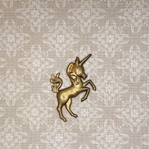 May include: A golden-colored unicorn figurine with a long horn and flowing mane. The unicorn is posed rearing up on its hind legs. The background is a patterned beige fabric.