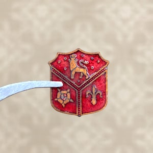 May include: A small, red and gold heraldic crest with a lion, rose, and fleur-de-lis design. The crest is held by tweezers, showcasing its intricate details and vintage aesthetic. The shield-shaped emblem is likely a decorative element.