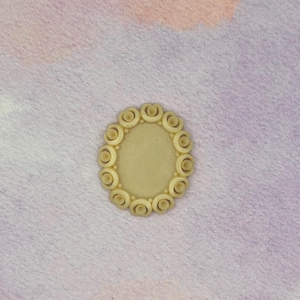 May include: An oval-shaped cream-colored cabochon featuring a smooth, flat center surrounded by a decorative border of small, raised circular rings. The piece has a uniform matte finish and a consistent light beige tone throughout its structure.