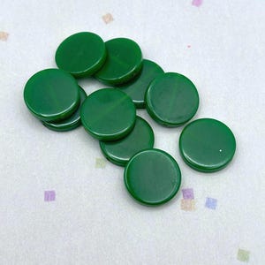 May include: A collection of glossy, circular, emerald green game pieces scattered on a white surface. The pieces are uniform in size and shape, with a smooth, reflective finish. The background has small, colorful, square patterns.