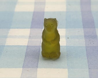 May include: A translucent, yellowish-green gummy bear candy in the shape of a bear. The gummy bear is standing upright with its arms folded in front. The candy is on a blue and white checkered surface.
