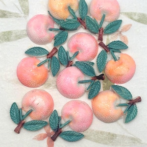 May include: Overhead shot of several peach-shaped ornaments. The ornaments are pink and orange with a textured surface. Each peach has a green leaf and brown stem attached. The background is a light-colored surface with a floral pattern.