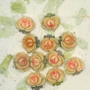 May include: A collection of eleven small, artificial rose blossoms. The roses are a pale yellow with pink centers and green leaves. The roses are arranged on a textured, off-white surface with green leaf accents.