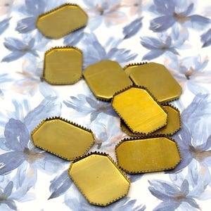 May include: A collection of gold-toned metal settings with a rectangular shape and notched, serrated edges. These flat, blank components feature a smooth, brushed surface finish and are designed for jewelry making or decorative craft projects.