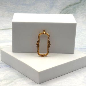 Peut inclure: Un cadre de pendentif en métal doré présentant une forme rectangulaire allongée avec des détails en filigrane décoratifs sur les côtés et une petite boucle circulaire au sommet pour la fixation, avec un design central ouvert.