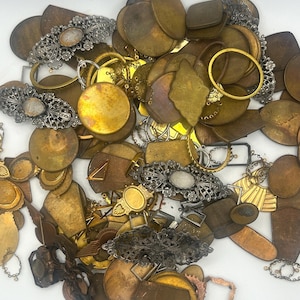 May include: A collection of vintage brass and silver-toned metal jewelry findings and components. The assortment includes various shapes like circles, ovals, and irregular forms, with some pieces featuring ornate filigree and decorative elements. The pieces vary in color from gold to bronze.