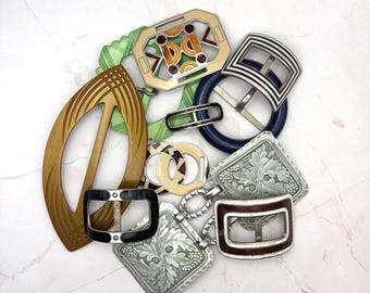 Puede incluir: Una colección de hebillas de cinturón variadas en diferentes formas y materiales, incluyendo una gran hebilla ovalada dorada, un marco geométrico verde, una hebilla circular azul oscuro, cuadrados plateados con relieve floral y una hebilla rectangular negra.