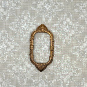 May include: An elongated, oval-shaped metal frame with a metallic bronze finish. The frame features intricate, raised decorative detailing along its entire border, including ornate, shell-like motifs at both the top and bottom center points.