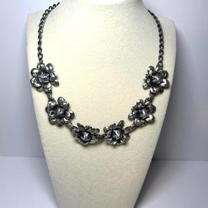 May include: A silver-coloured necklace with a floral design. The necklace is made up of several flower-shaped pendants linked together by a chain. The necklace is displayed on a white mannequin bust. The flowers have textured petals.