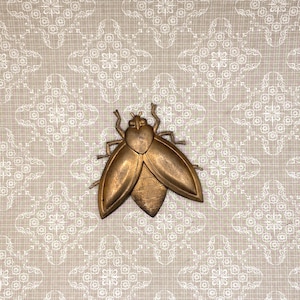 Puede incluir: Un insecto decorativo de metal dorado con alas y cuerpo detallados. El insecto está centrado en un fondo beige con un patrón floral blanco. El insecto es un elemento decorativo.