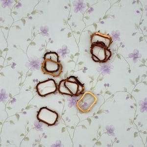 May include: A collection of seven small, rectangular, metallic frames with a copper-toned finish. Each frame features four tiny, pointed prongs extending from the corners, designed for securing small items within the open center of the structure.