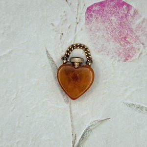 May include: A heart-shaped amber-colored pendant with a marbled texture. The pendant features a metallic gold-toned screw-top cap and a small attached gold-toned chain link at the top, allowing it to be suspended as a decorative charm or jewelry piece.