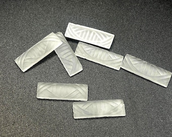 May include: Seven rectangular, frosted glass beads with a textured, curved design. The beads are arranged on a dark gray surface. The glass has a translucent quality, allowing light to pass through.