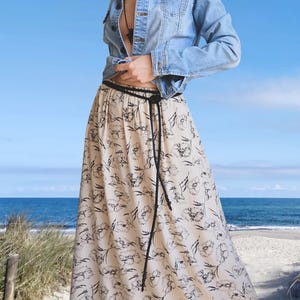 May include: A light blue denim jacket with a button-up front paired with a long, cream-coloured skirt featuring a black floral print. The skirt is cinched at the waist with a thin, black braided cord belt that hangs down the front of the garment.
