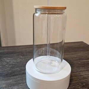 Puede incluir: Un vaso de vidrio transparente con tapa de madera y una pajita. El vaso est&aacute; sobre un pedestal blanco.