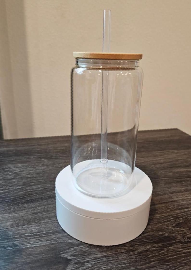 Puede incluir: Un vaso de vidrio transparente con tapa de madera y una pajita. El vaso est&aacute; sobre un pedestal blanco.