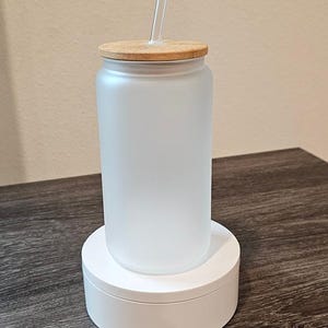 Puede incluir: Un vaso de vidrio esmerilado con tapa de bamb&uacute; y una pajita transparente. El vaso est&aacute; sobre un pedestal blanco.