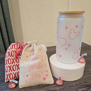 Puede incluir: Dos peque&ntilde;as bolsas de arpillera con corazones rosas y el texto "Happy Valentine's Day" y "XOXO" impreso en ellas. Un vaso de vidrio esmerilado con un dise&ntilde;o de Mickey Mouse rosa y una tapa de madera. El vaso est&aacute; sobre un pedestal blanco. Hay besos de chocolate rosa sobre la mesa.
