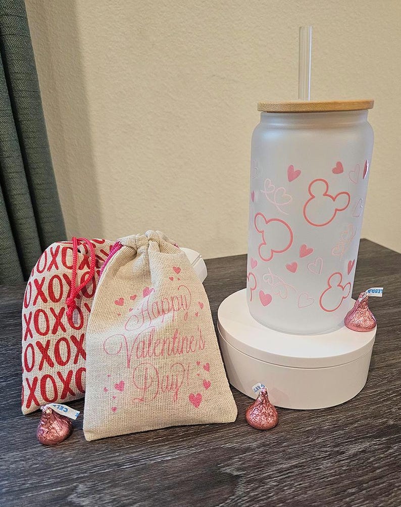 Puede incluir: Dos peque&ntilde;as bolsas de arpillera con corazones rosas y el texto "Happy Valentine's Day" y "XOXO" impreso en ellas. Un vaso de vidrio esmerilado con un dise&ntilde;o de Mickey Mouse rosa y una tapa de madera. El vaso est&aacute; sobre un pedestal blanco. Hay besos de chocolate rosa sobre la mesa.