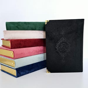 Peut inclure: Une pile de cinq livres recouverts de velours avec des accents dorés. Les livres sont de différentes couleurs : vert, rose, rouge, rose, bleu clair et bleu marine. Le livre du dessus est noir avec des accents dorés et de la calligraphie arabe.