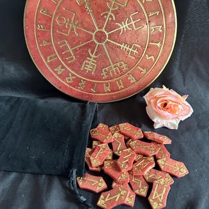 Op de afbeelding: Een set van 24 runen, een rode fluwelen zak en een rode ronde steen met gouden runen en een kompasontwerp.