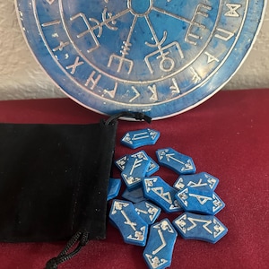 Op de afbeelding: Een blauwe fluwelen zak met een trekkoord, die een set blauwe runenstenen met zilveren markeringen bevat. De runen zijn gerangschikt op een rood oppervlak, met een blauw en zilver rond object op de achtergrond.