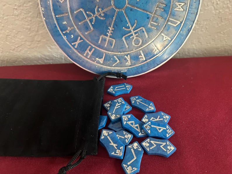 Op de afbeelding: Een blauwe fluwelen zak met een trekkoord, die een set blauwe runenstenen met zilveren markeringen bevat. De runen zijn gerangschikt op een rood oppervlak, met een blauw en zilver rond object op de achtergrond.