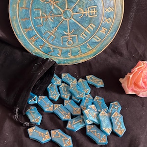 Op de afbeelding: Een set blauwe en gouden runenstenen met gouden belettering op elke steen. De stenen zijn verspreid over een zwart oppervlak. Een blauw en gouden rond houten bord met runen is op de achtergrond.